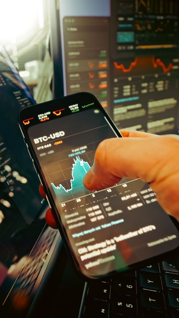 about-us-01 A hand holds a smartphone displaying BTC-USD chart amidst digital trading screens, highlighting crypto finance.